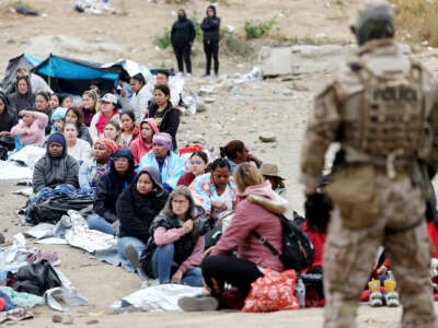 People sit in a line as they're watched by an armed and uniformed border patrol agent