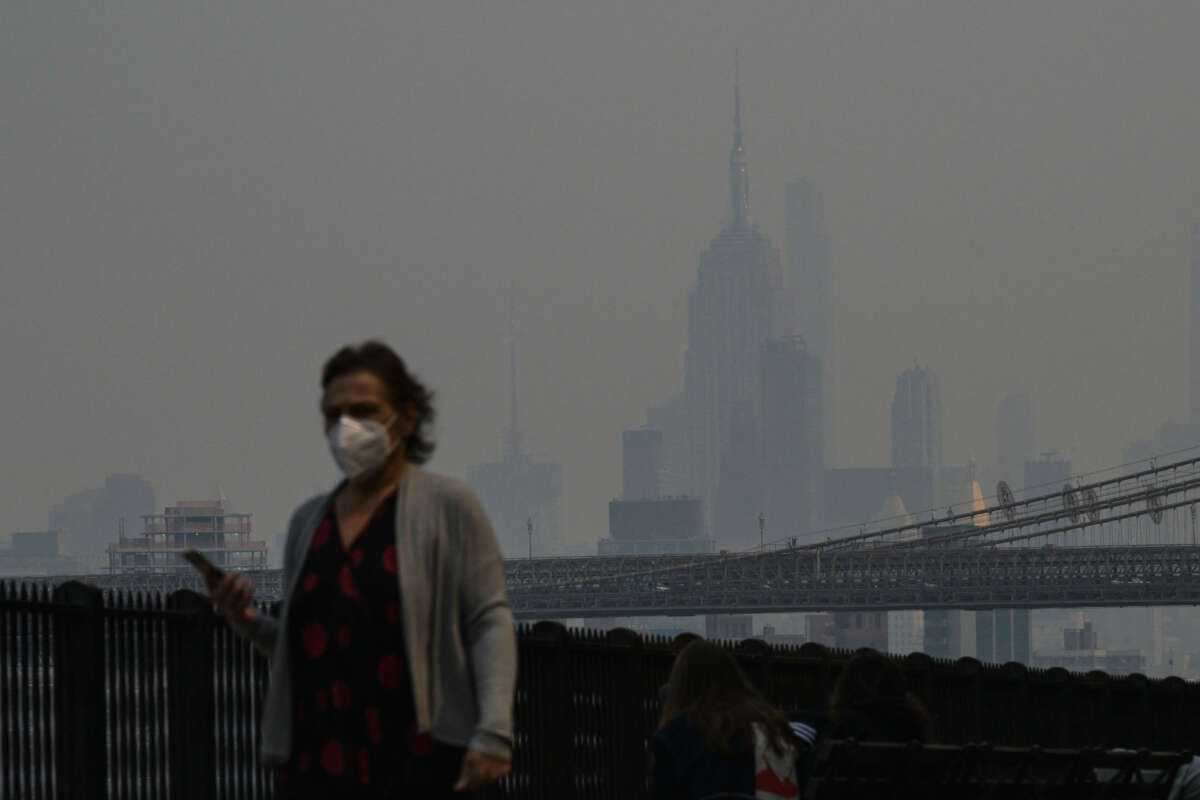 A man in a mask walks by a fence, beyond which the smoke-hazed skyline of New York City can be seen