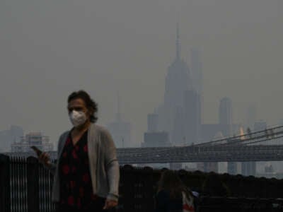 A man in a mask walks by a fence, beyond which the smoke-hazed skyline of New York City can be seen