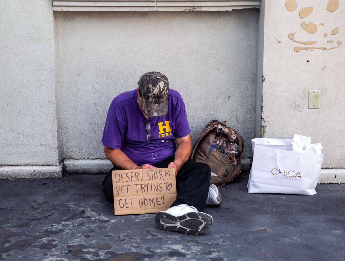 a begging man sits on the sidwalk behind a cardboard sign reading "DESERT STORM VET TRYING TO GET HOME!!"