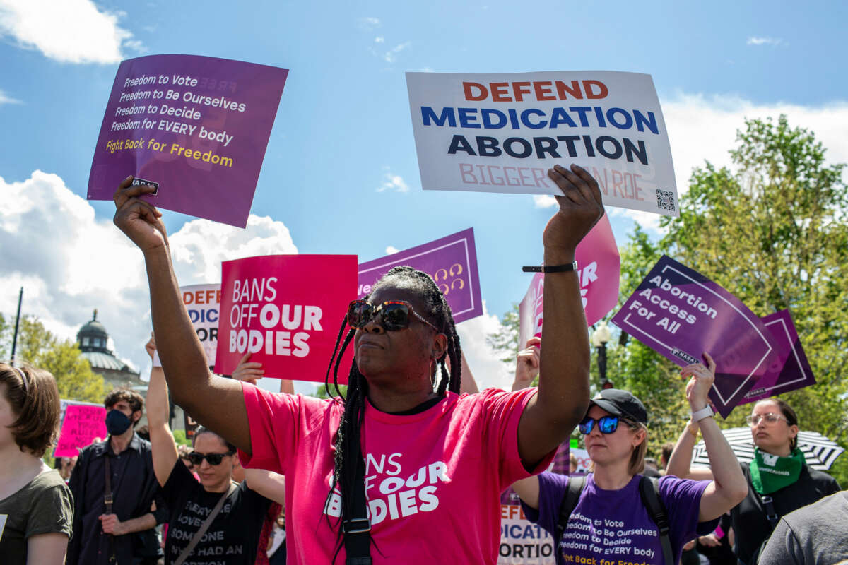 A Black woman holds signs reading "PROTECT MEDICATION ABORTION" and "Freedom to vote; freedom to be ourselves; freedom to decide; freedom for EVERYBODY; Fight back for freedom" during an outdoor protest