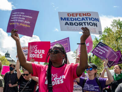 A Black woman holds signs reading "PROTECT MEDICATION ABORTION" and "Freedom to vote; freedom to be ourselves; freedom to decide; freedom for EVERYBODY; Fight back for freedom" during an outdoor protest