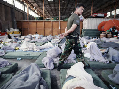 A man in military uniform patrolls refugees as they sleep on the floor of a warehouse