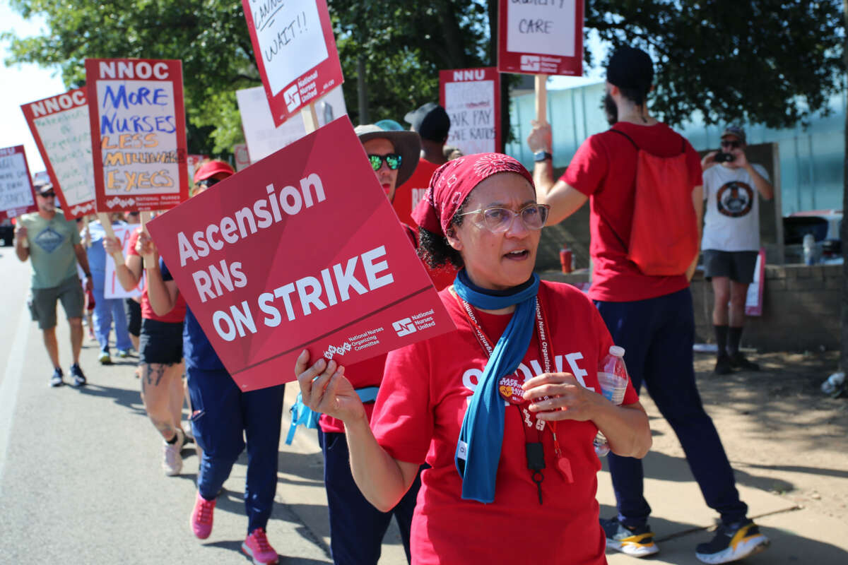 A protester dressed in red holds a sign reading "Ascension RNs ON STRIKE" alongside others, also holding signs and wearing red