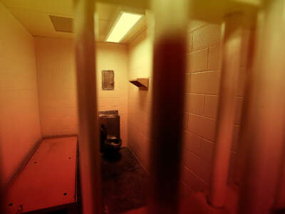 A photo of the interior of a jail cell, that has been edited with a red and yellow overlay to imply heat. It's about the size of a parking space, and contains a toilet, a twin bedframe with no, pillow or blanket, and nothing else.