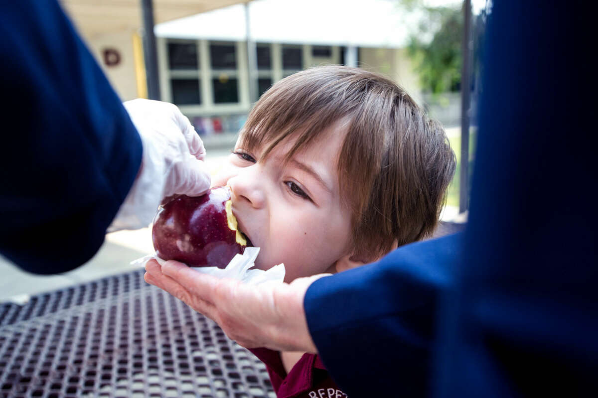 A boy receives help eating an apple from an adult wearing gloves