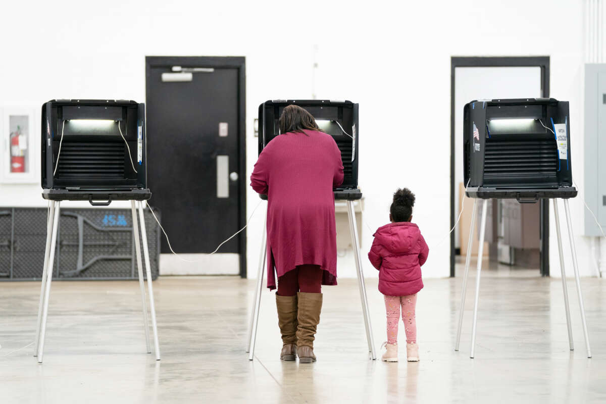 A woman in pink votes at a booth as her young daughter, wearing a coat in the same shade of pink as her mother, stands beside her