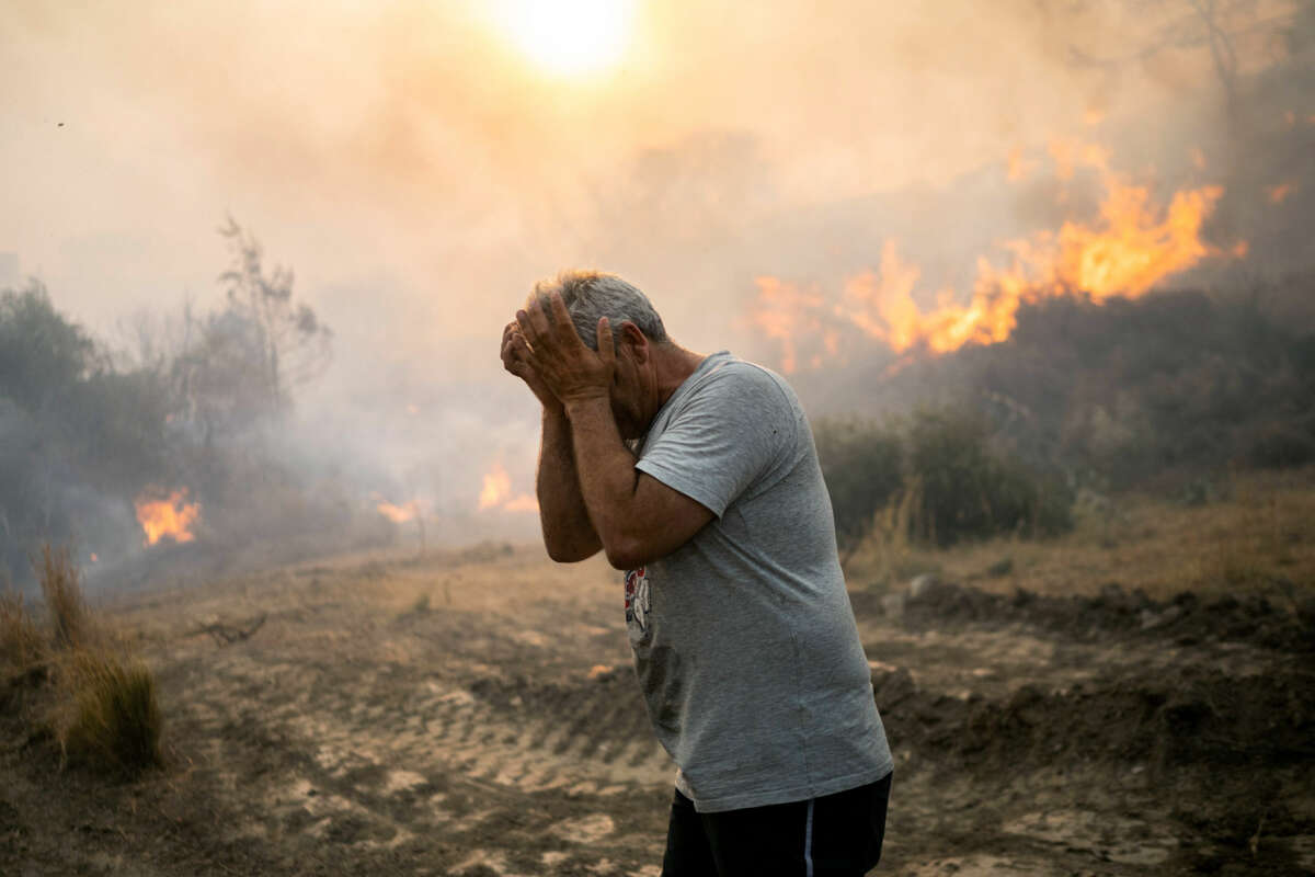 A man rubs his eyes as the landscape behind him is consumed in fire and smoke