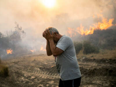 A man rubs his eyes as the landscape behind him is consumed in fire and smoke