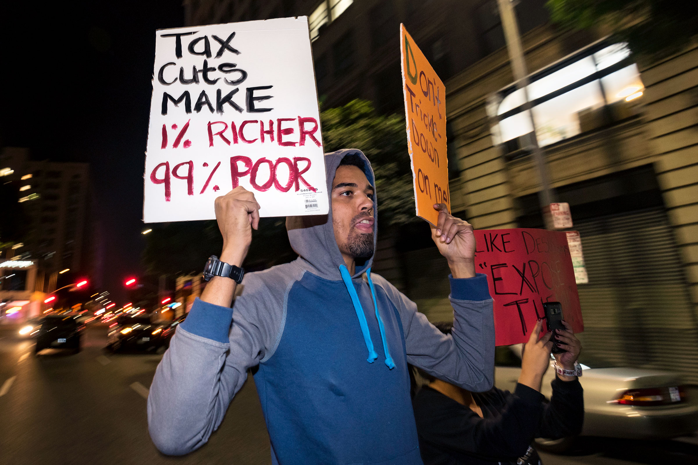 A protester holds a sign reading "TAX CUTS MAKE 1% RICHER, 99% POOR" during an outdoor protest