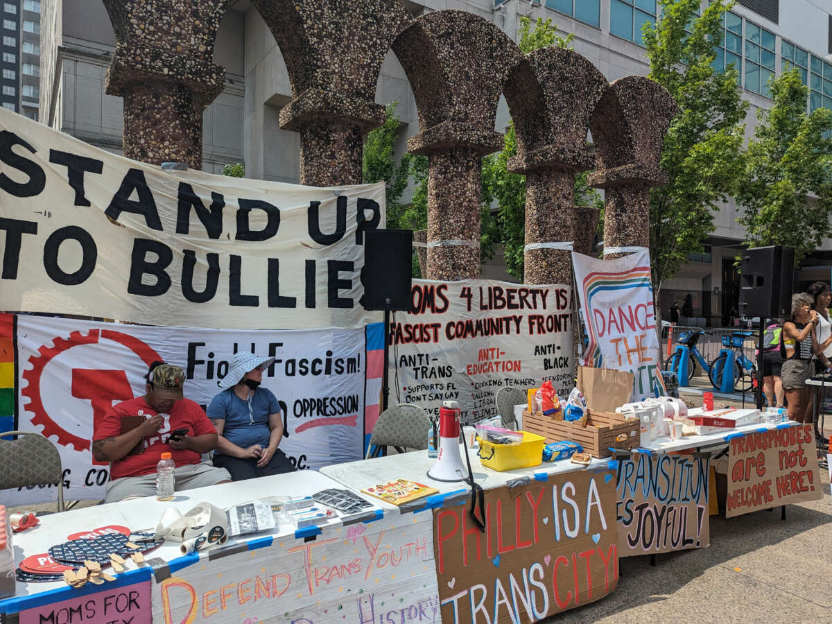 Tables with snacks, fans, COVID rapid tests, water and coffee are surrounded by signs and banners with slogans like "Stand up to Bullies" and "Transition is Joyful" at the protests on July 1, 2023.