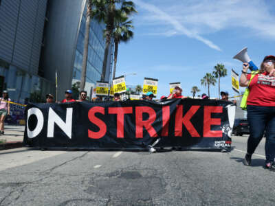 People march behind a banner reading "ON STRIKE" as a person chants on a megaphone