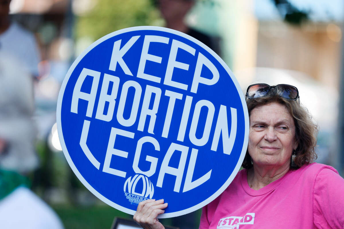 An older woman standing outdoors holds a sign reading "KEEP ABORTION LEGAL"