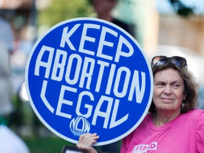 An older woman standing outdoors holds a sign reading "KEEP ABORTION LEGAL"