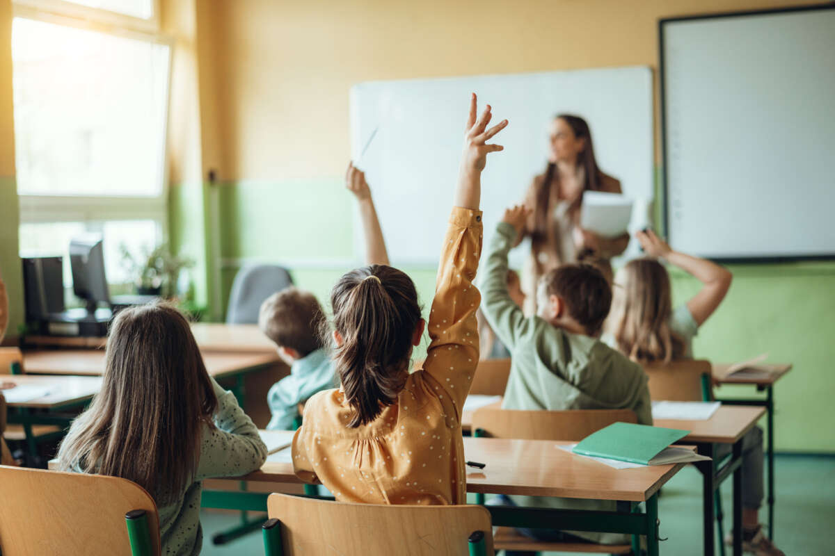 Students raise hands in class, teacher out of focus in background