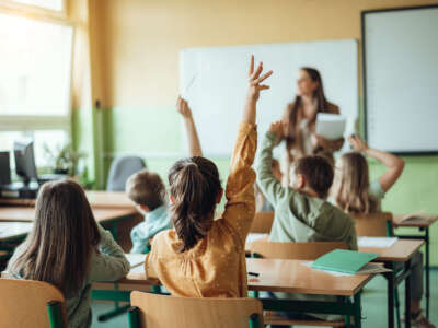 Students raise hands in class, teacher out of focus in background