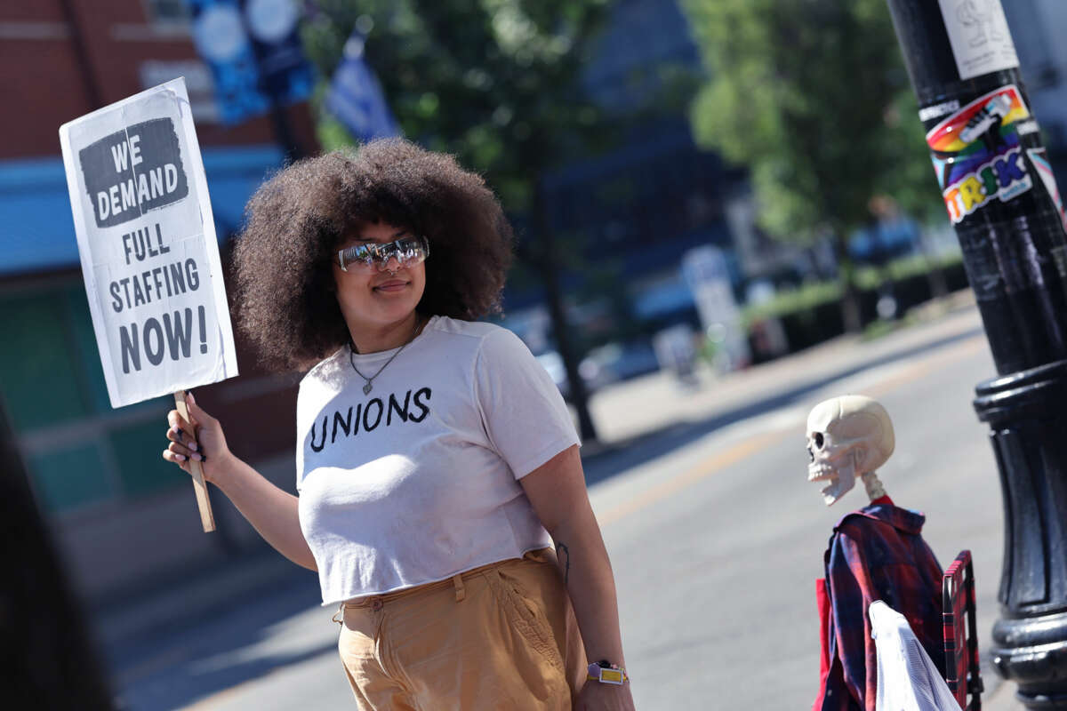 A protester wearing a shirt that reads "UNIONS" holds a sign a sign reading "WE DEMAND FULL STAFFING NOW" during an outdoor protest