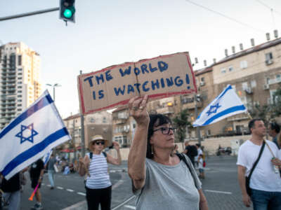 A woman, flanked by protesters behind her carrying the Israeli flag, holds a sign reading "THE WORLD IS WATCHING"
