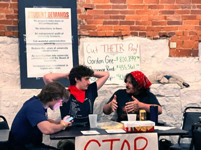 Three students - one wearing a black shirt, and looking at his phone; one wearing a red bandanna around his neck, with his arms behind his head casually; and one wearing a red bandana on his head, speaking to the student wearing the bandana around his neck. All three are seated at a table in front of banners reading "CUT THEIR PAY" with the six-figure salaries of the university's executives listed below, alongside a printed list of the student body's demands.