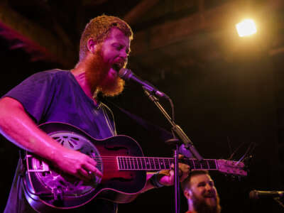 Oliver Anthony is a bearded white man standing in front of microphone playing a guitar bathed in neon light.