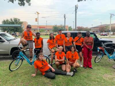 10 people, 9 of whom are wearing matching orange shirts, and the last of whom is not, pose for a photo with bicycles