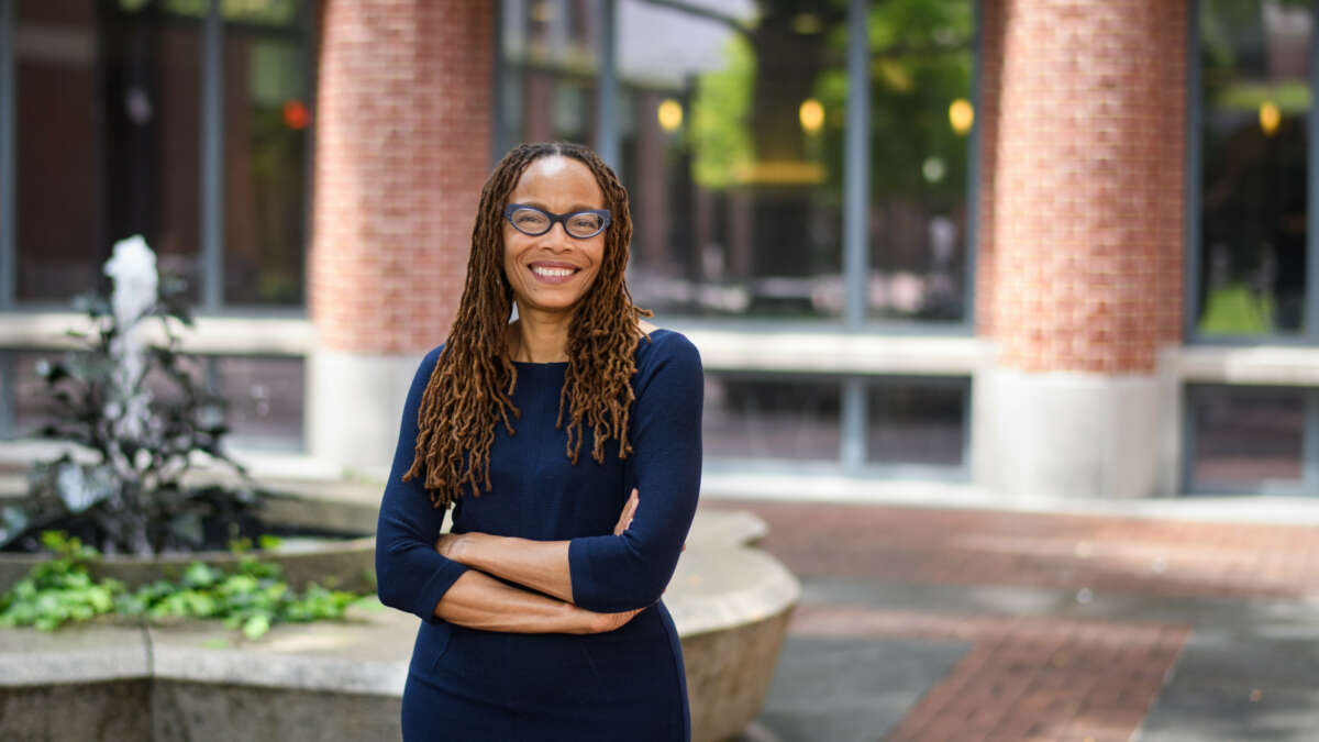 Dorothy Roberts, a light-skinned Black woman with wavy locs and glasses, smiles and crosses her arms in front of a college campus