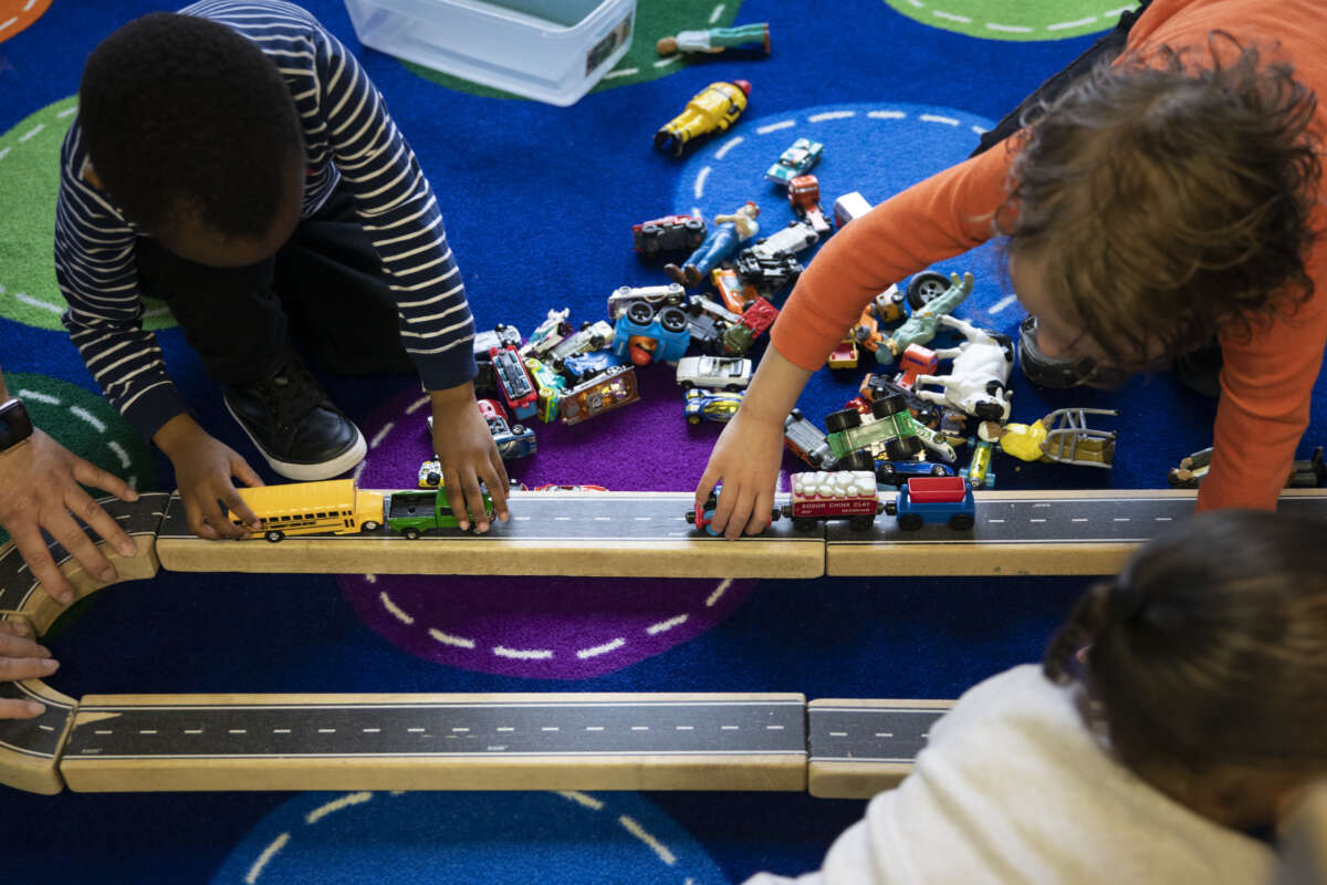 Two children play together with toy cars along a wooden track in a classroom