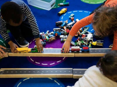 Two children play together with toy cars along a wooden track in a classroom