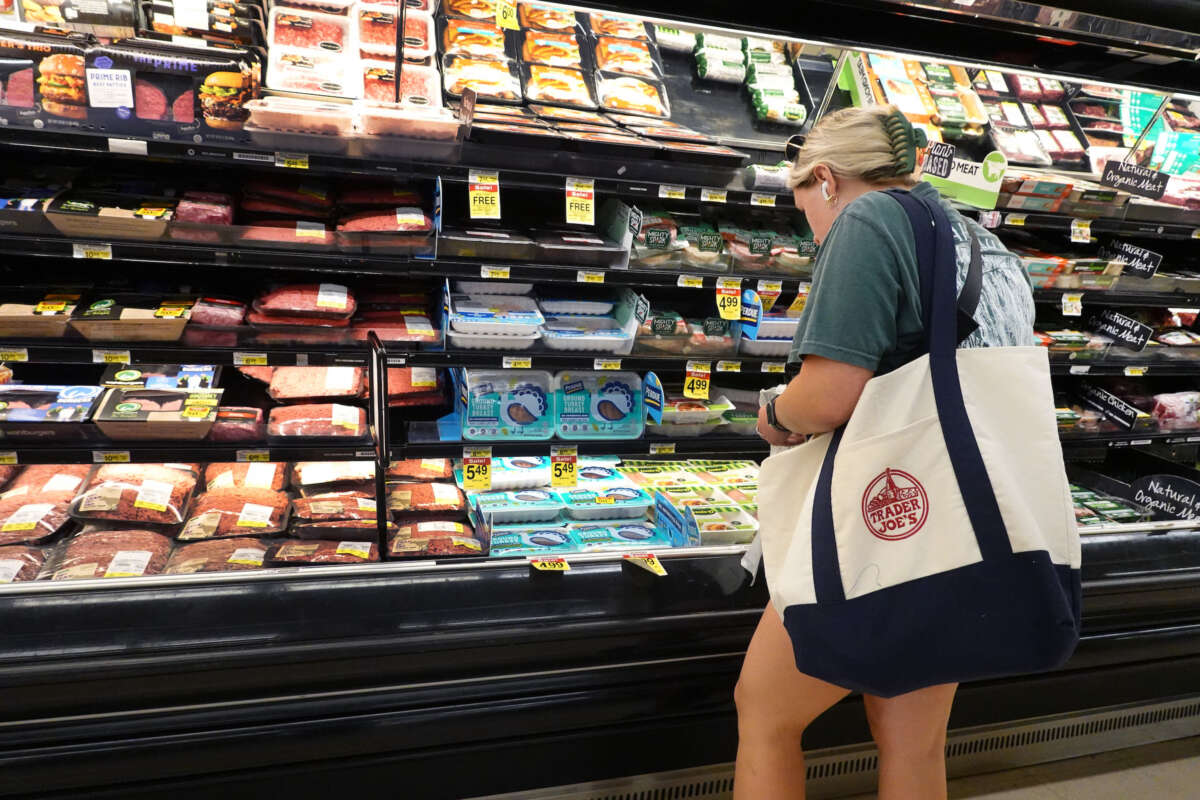 A blonde woman shops for groceries in the meat section of a grocery store.