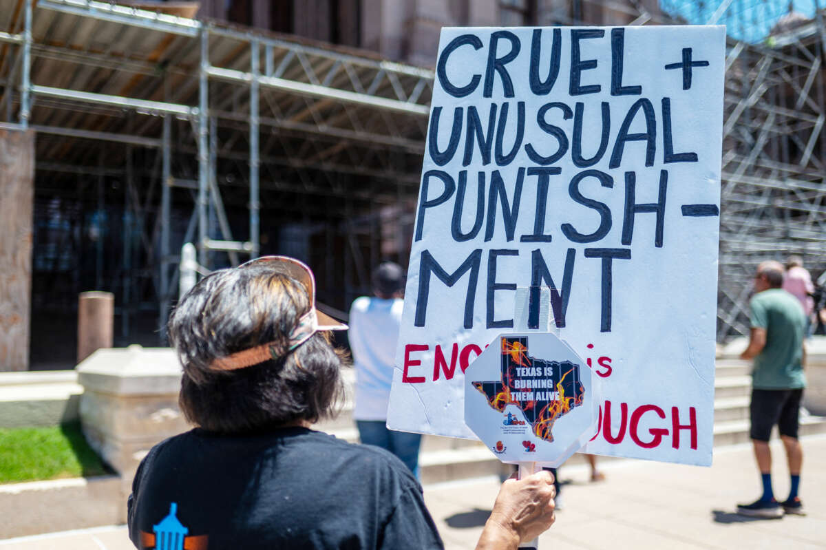 A protester, their back facing the viewer, holds a sign reading "CRUEL AND UNUSUAL PUNISHMENT; ENOUGH IS ENOUGH" during an outdoor protest