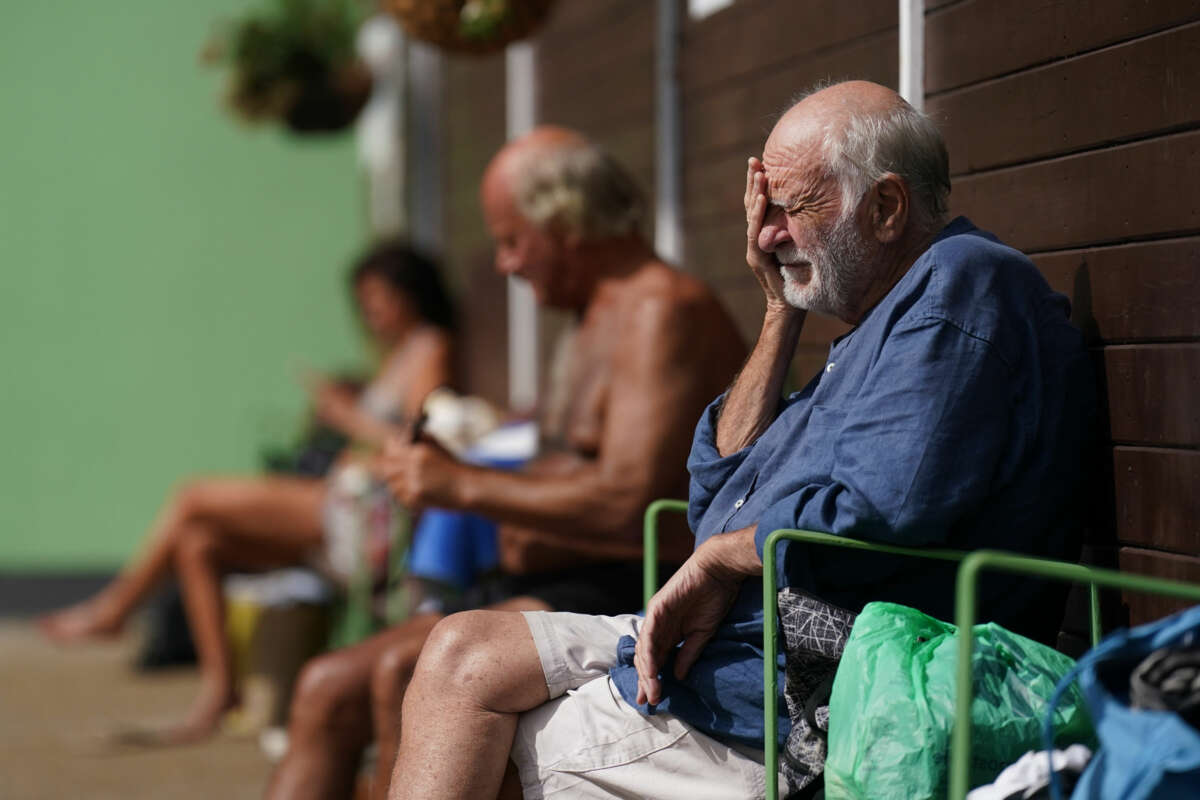 An elderly man sits against a wall and rests his face in his hands