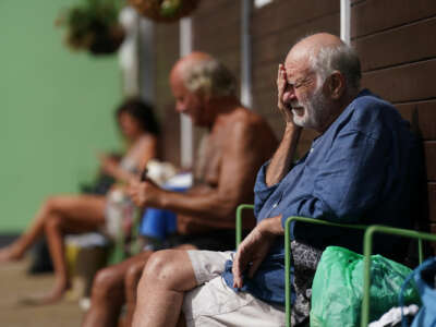 An elderly man sits against a wall and rests his face in his hands