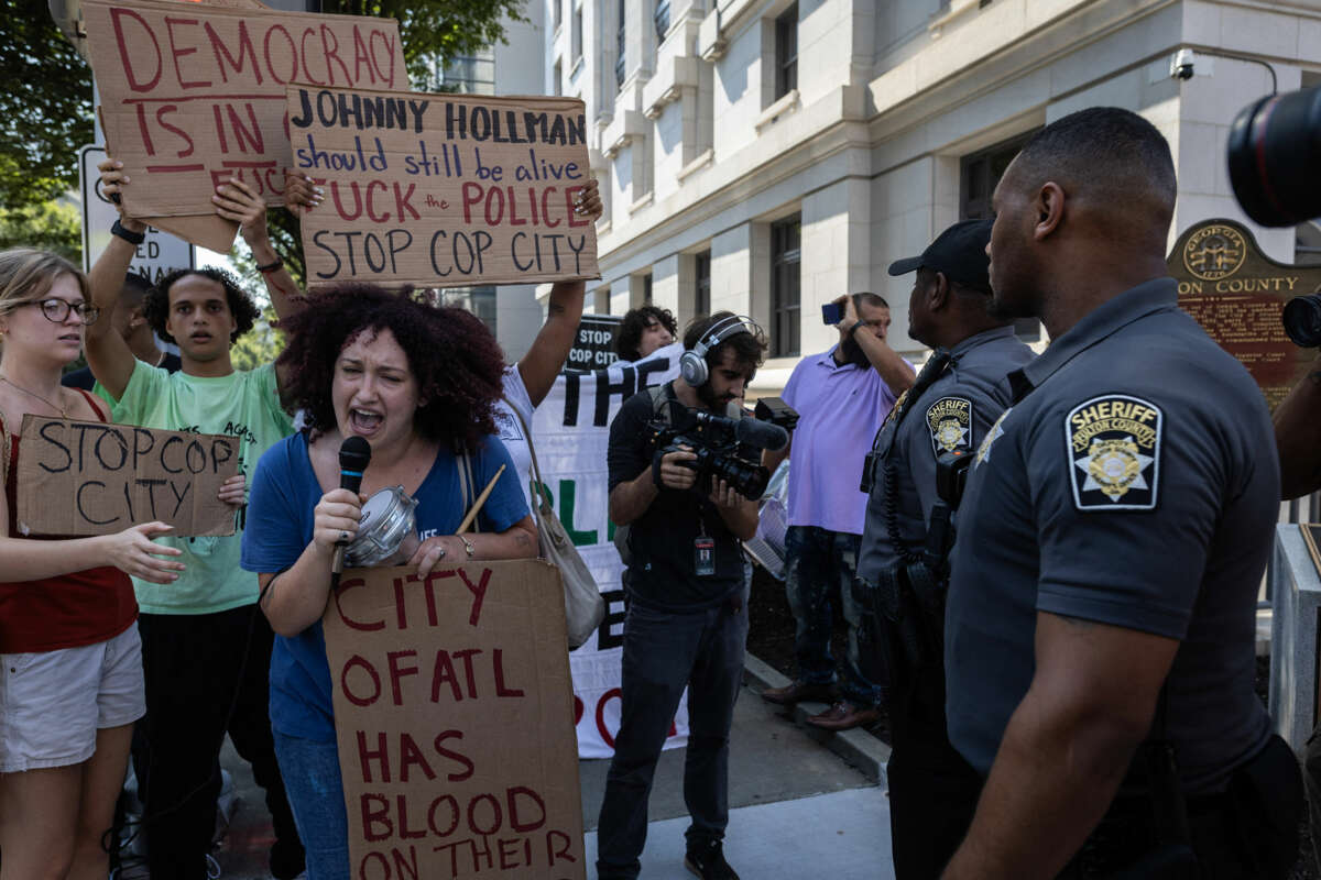 A person displays a sign reading "Johnny Hollman should still be alive; *uck the police; stop cop city" behind another protester, holding a sign reading "THE CITY OF ATL HAS BLOOD ON THEIR HANDS" while speaking into a microphone