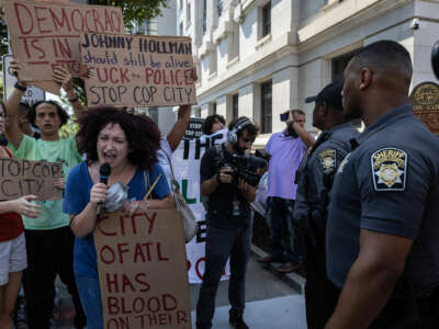 A person displays a sign reading "Johnny Hollman should still be alive; *uck the police; stop cop city" behind another protester, holding a sign reading "THE CITY OF ATL HAS BLOOD ON THEIR HANDS" while speaking into a microphone