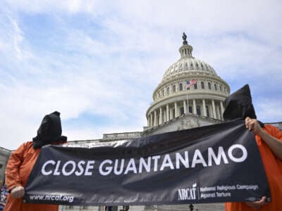 Two demonstrators in orange jumpsuits wearing black hoods carry a sign saying "Close Guantanamo" in front of the Capitol.