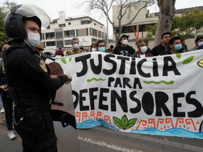 People stand behind a banner reading "JUSTICA PARA DEFENSORES" as police in riot gear stand in front of them