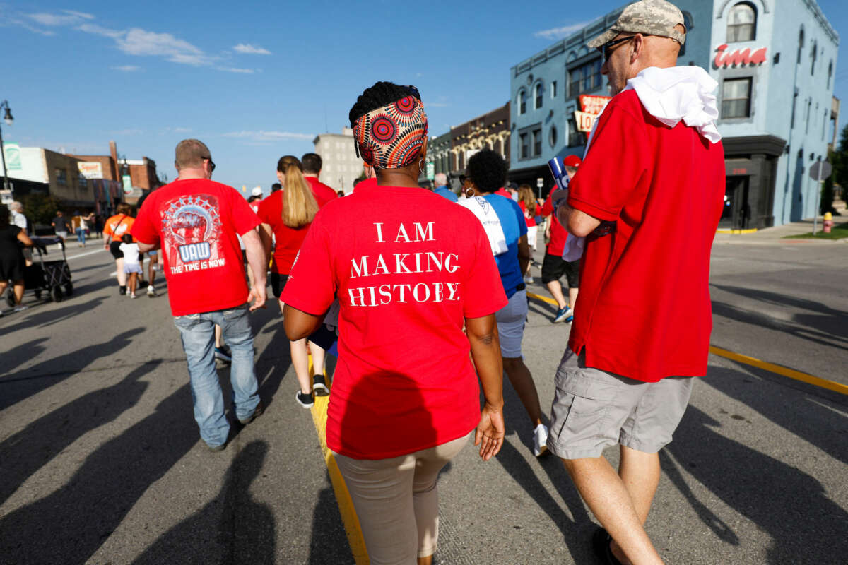 a woman wearing a headwrap and a shirt that reads "I AM MAKING HISTORY" walks with other people during an outdoor strike