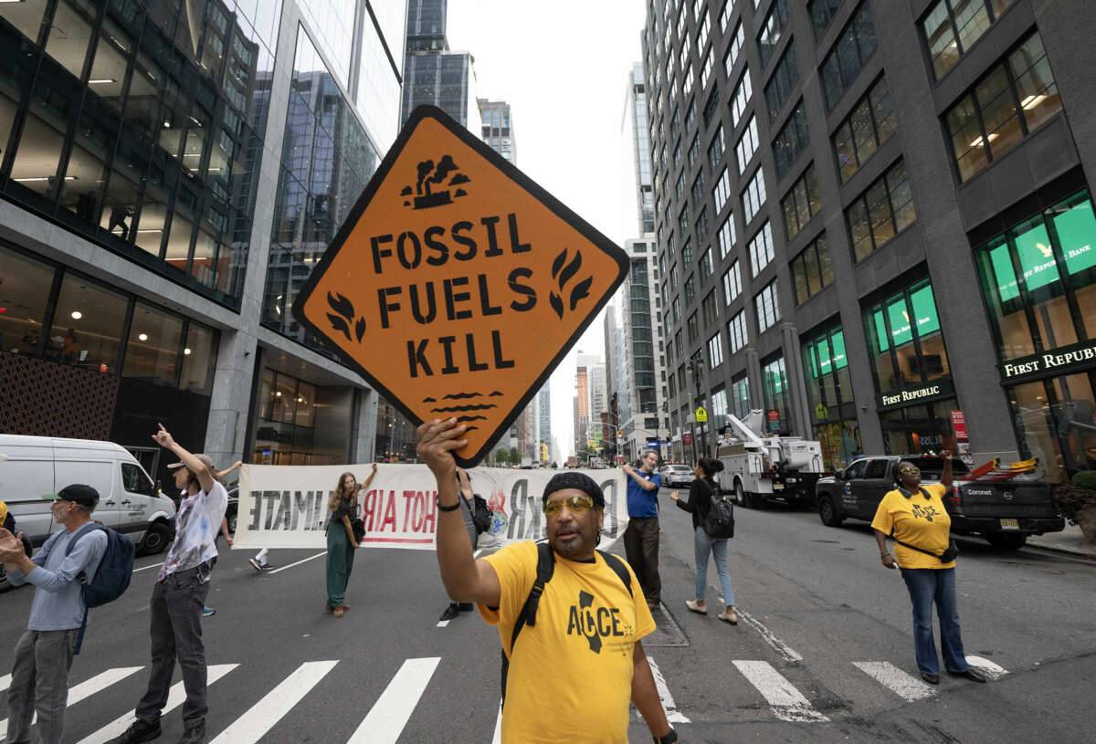 A protester holds a fake road sign reading "FOSSIL FUEL KILLS" during an outdoor street protest