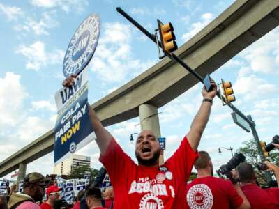 A man raises a sign with arms outstretched that reads "UAW Stand Up COLA and Fair Pay Now"