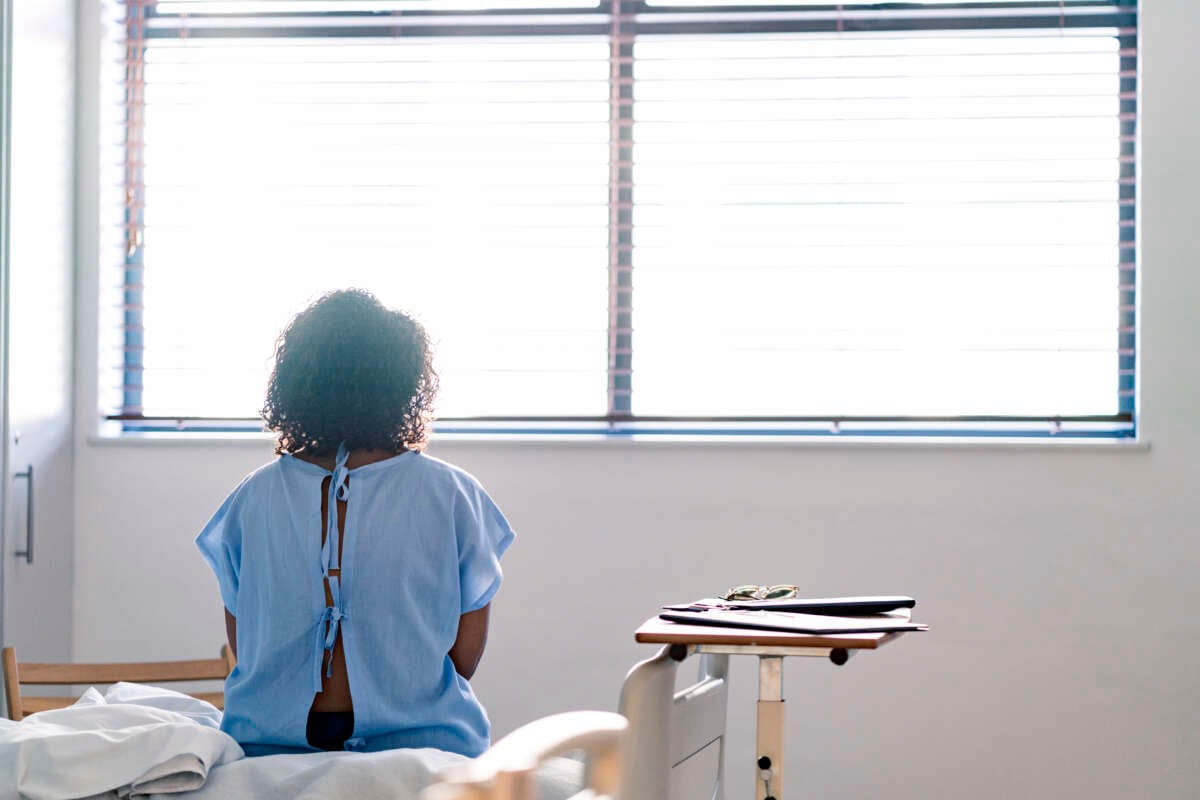 Young Black patient wearing a hospital gown sits on a table and looks out the window