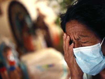 An elderly patient in a medical mask wearily rests her face against her palm
