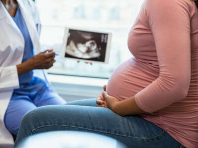 A photo with the focus on the unrecognizable pregnant woman in the foreground as the unrecognizable doctor shows her an ultrasound on a digital tablet in the background.