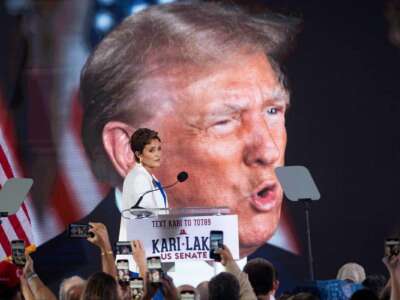 Kari Lake walks in front of a banner of Donald Trump's face