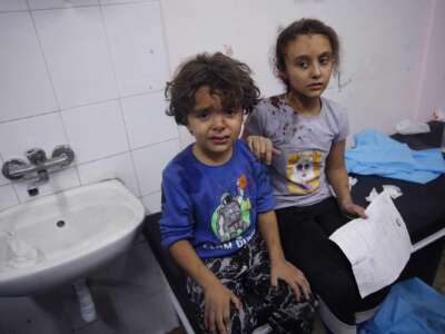 Two injured Palestinian children, one looking directly into the camera, sit on a bench inside of a hospital