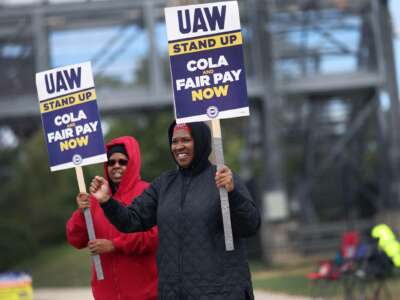 Two strikers stand outside holding signs reading "UAW STAND UP: COLA AND FAIR PAY NOW" during an outdoor rally