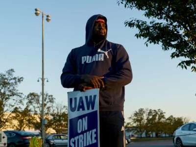 A man in a hoodie stands behind his "UAW ON STRIKE" sign as he gazes beyond the view of the camera