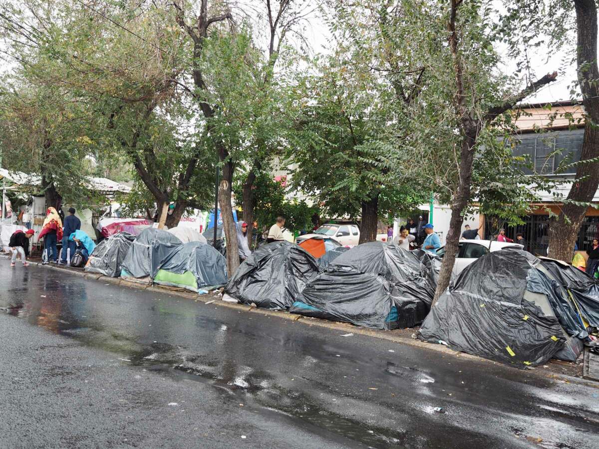 Tents covered in black plastic line a paved road as people walk around