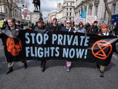 People march behind a banner reading "STOP PRIVATE FLIGHTS NOW" during an outdoor demonstration