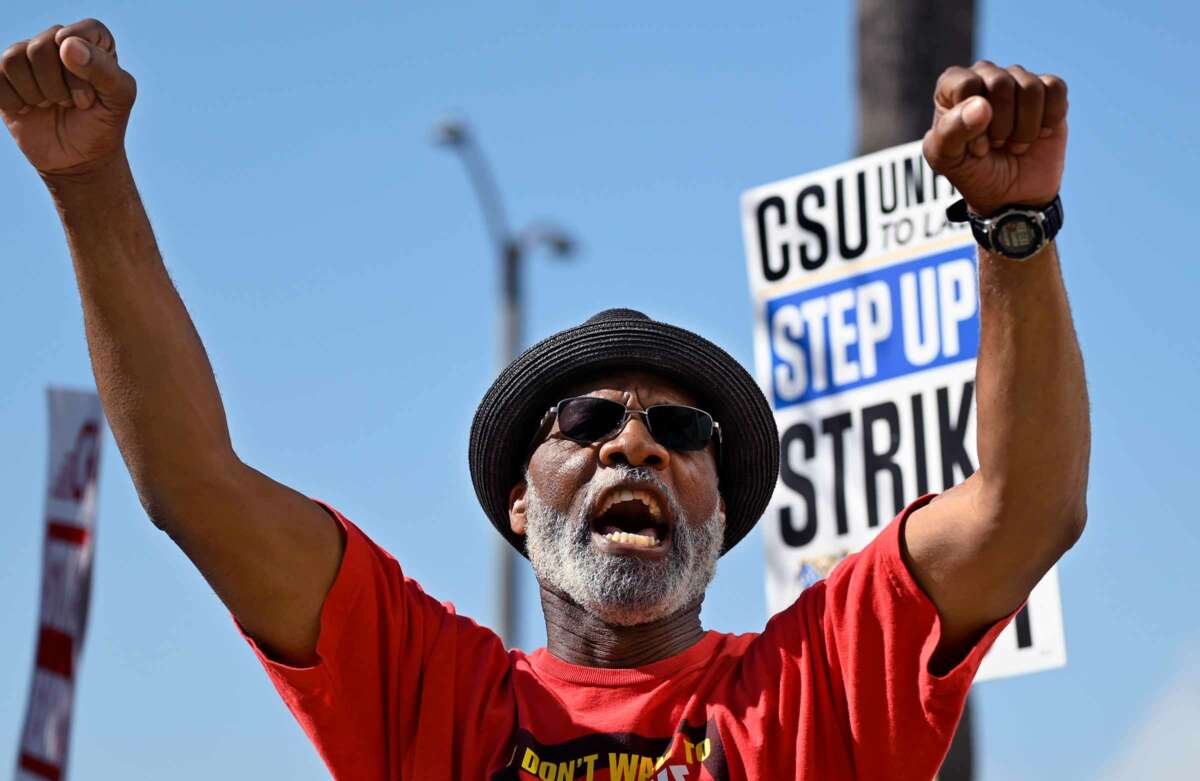 A man raises both fists and chants during an outdoor demonstration as a sign reading "CSU: STEP UP STRIKE" is partially visible behind him