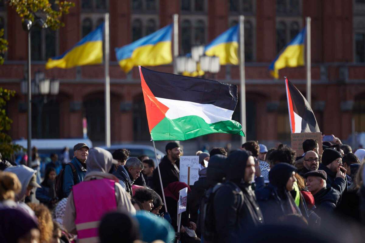 A person holds a Palestinian flag while standing in a public square flying Ukrainian flags during an outdoor rally